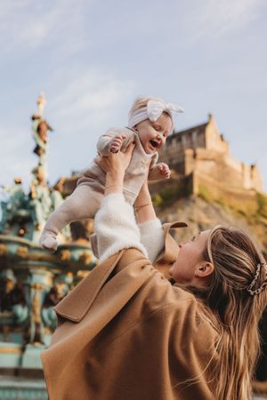 A parent in a brown coat lifts a baby wearing white clothing against a sunny outdoor backdrop with buildings.