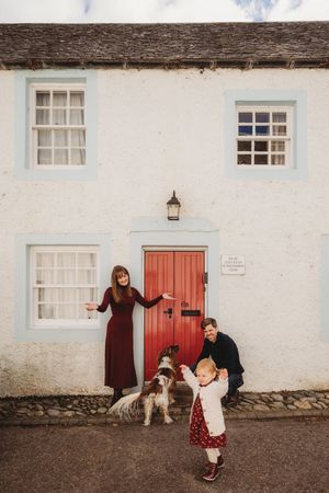 A family photoshoot outside a white cottage with red doors, featuring playful interactions with their dog.