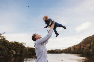 A parent lifts a small child high into the air against a scenic autumn lake backdrop.
