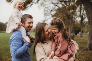 Happy family of four enjoying autumn day in park, parents holding two young daughters while laughing together outdoors.