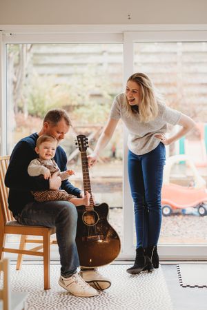 A happy family with a man holding a baby and playing guitar while a woman laughs beside them indoors.