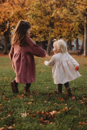 Two young girls in dresses hold hands playing in a park surrounded by colorful autumn foliage and fallen leaves.