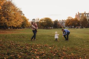 Family playing in an autumn park with golden trees, parents and toddler enjoying outdoor fun on green grass.