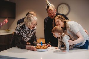 Family gathering around a table celebrating a baby's first birthday with a small cake and candle indoors.