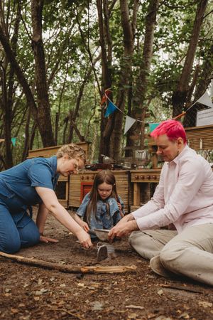 Family of three arranging sticks together outdoors in a woodland play area with bunting and rustic wooden structures.