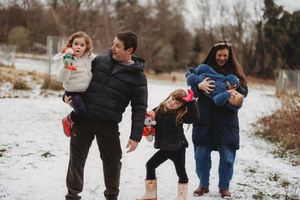 Family of five playing in light snow outdoors, parents holding toddlers while young girl dances between them.