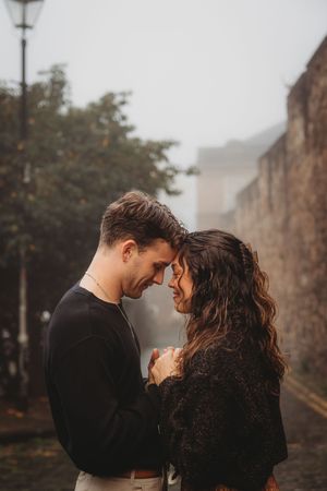 Romantic couple sharing an intimate moment on a misty autumn street with stone walls and lamp post in background.