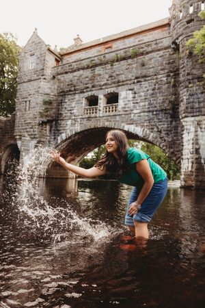 Young woman in green top splashing water in a river, with a historic stone arch bridge in the background.