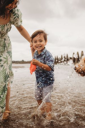 A child splashes and plays in the shallow ocean water on a cloudy beach day while holding someone's hand.