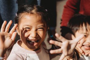 Joyful young Asian girl laughing with hands raised, sharing a happy moment with friends in warm natural light.