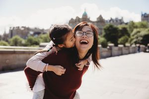 A young girl gives a piggyback ride kiss to a laughing woman wearing glasses outdoors in a sunny city setting.