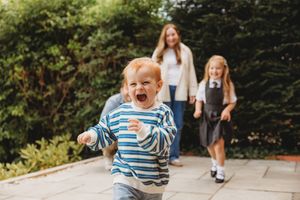 Toddler boy running and laughing outdoors on a patio with a woman and young girl in school uniform in the background.