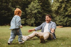 Father sitting on grass laughing with arms open as toddler with red hair walks toward him in park.