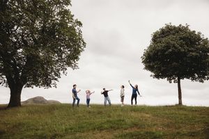Five people playing and dancing on a grassy hill between two trees under an overcast sky.