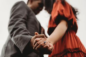 Couple holding hands with engagement ring visible, woman in red dress and man in gray suit, romantic outdoor photo.