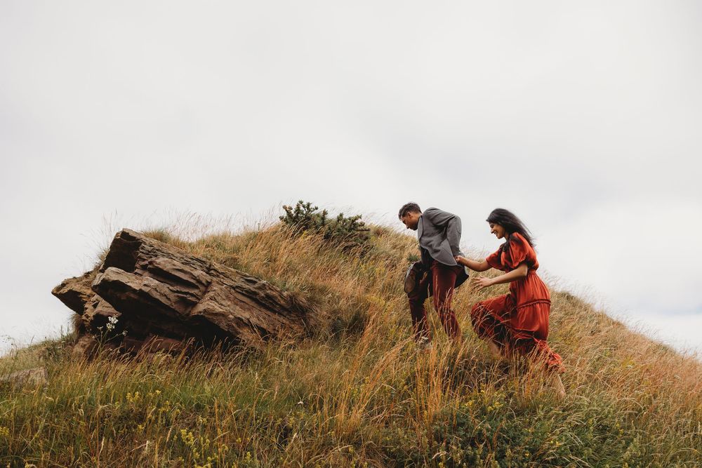 Couple in red dress and gray jacket walking through tall grass on a hillside near a large rock on an overcast day.
