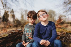 Two young boys sitting on a mossy log outdoors, one wearing glasses and smiling, surrounded by autumn trees.