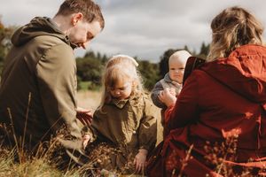 Family with two young children exploring nature in a field, parents crouching down with toddler and baby outdoors.