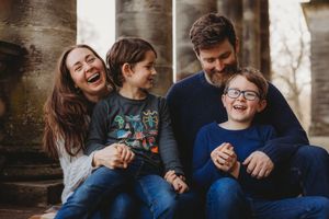Happy family of four laughing together outdoors, sitting on stone steps in casual clothing.