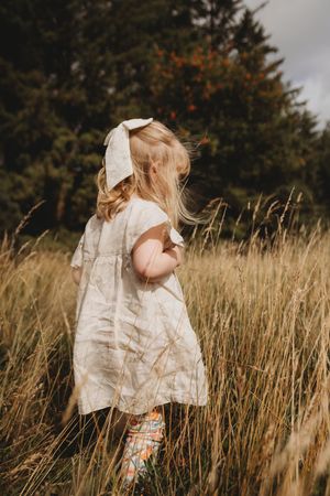 Young blonde girl in a white linen dress and bow walks through tall golden grass in a lush green field.