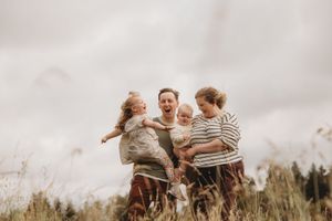 Happy family playing in a field, parents laughing and swinging children outdoors on a cloudy day.