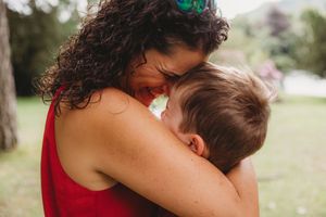 A mother in a red dress tenderly embraces her young child outdoors in a lush green park setting.