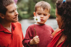 A young boy in a red sweater holds a daisy flower while standing between two adults outdoors.