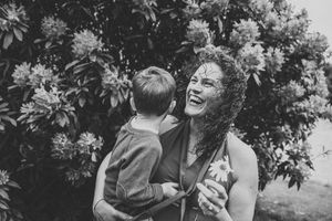 Black and white photo of a laughing woman holding a young child outdoors near blooming flowering shrubs.