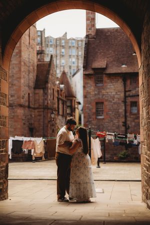 Couple sharing a romantic embrace under a stone archway in a historic Edinburgh courtyard with laundry lines in the background.