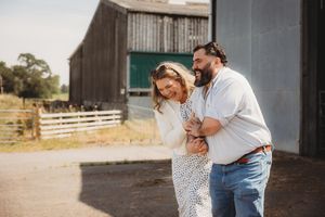Happy couple embracing and laughing together outside a rustic barn on a sunny farm day.