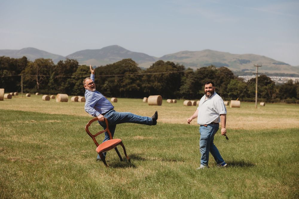 Two men laughing in a sunny field with hay bales and mountains, one balancing on a wooden chair outdoors.