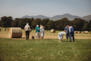 Group of people walking in a green field with hay bales and mountain backdrop on a sunny day.