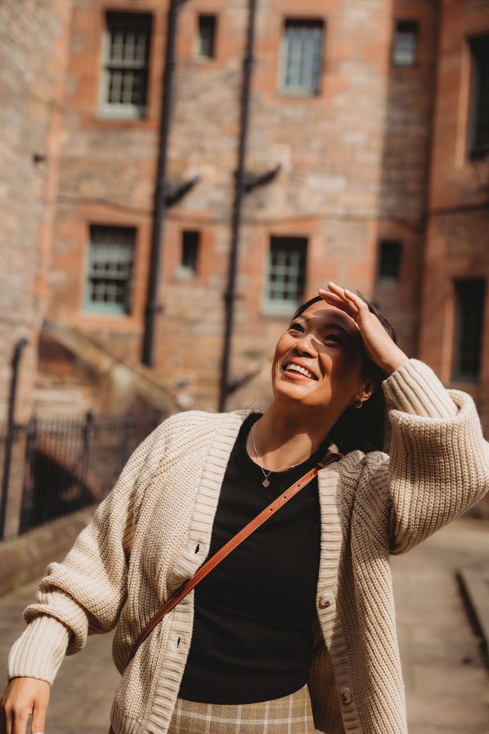 Smiling young woman in cream cardigan and crossbody bag shielding eyes from sun near historic brick building.