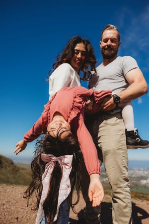 Happy family with parents holding their laughing child upside down on a sunny hilltop with blue sky.