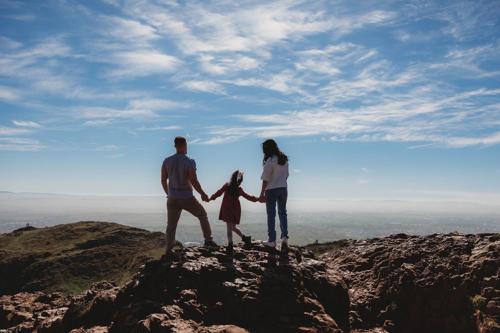 Family of three holding hands on rocky mountain summit with scenic blue sky and vast landscape view.