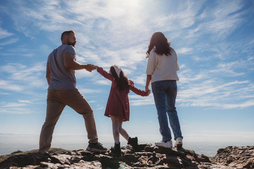 Family of three holding hands on rocky hilltop against blue sky, viewed from behind.