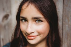 Close-up portrait of a young woman with freckles, brown hair, and a warm smile against a wooden background.