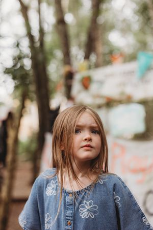 Young girl in blue floral dress standing outdoors among trees, looking thoughtfully into the distance.