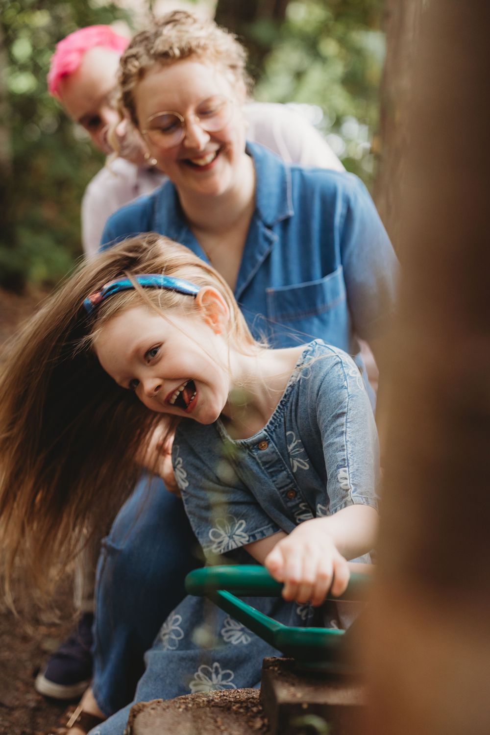 A laughing girl in denim holds a green handle outdoors while a smiling woman in blue watches from behind.