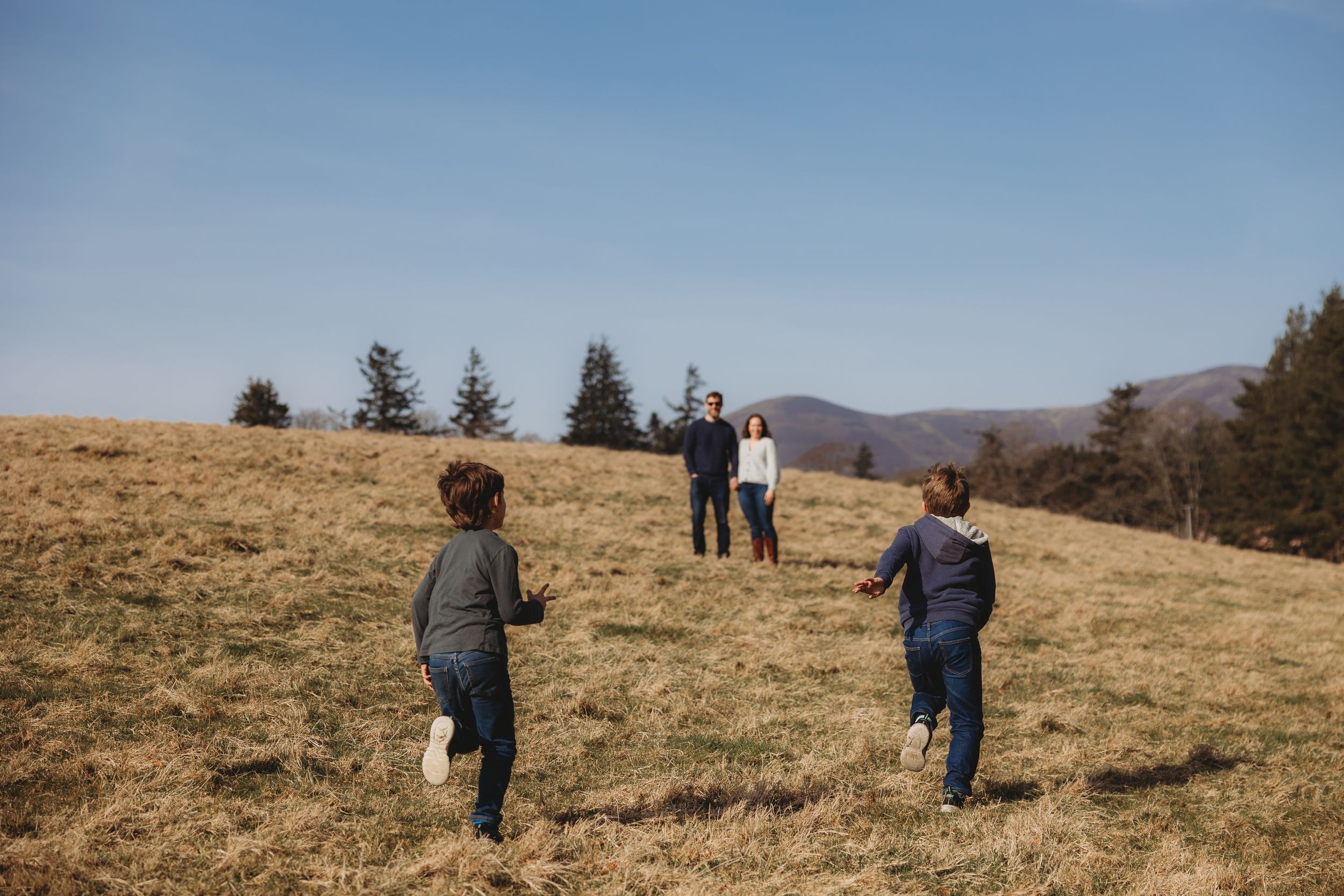 Two children running toward parents on a grassy hillside under a clear blue sky with mountains in the background.