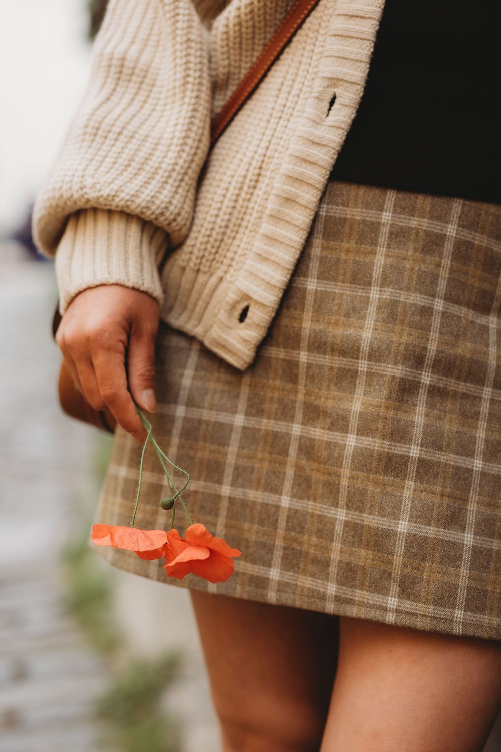 Woman in plaid mini skirt and cream knit cardigan holding a single red poppy flower.