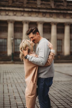 A couple embraces tenderly on a cobblestone plaza in romantic urban setting with classic architecture in background.