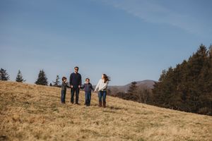 Family of four holding hands on a grassy hillside with pine trees and blue sky in the background.