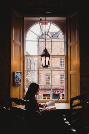 A silhouetted figure sits by an arched window in a dimly lit room with warm lighting and city buildings visible outside.