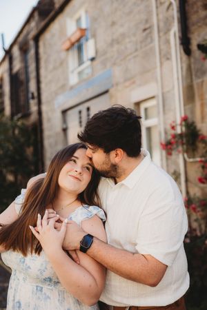 A couple embraces tenderly in front of a rustic stone building, man kissing woman's temple as she smiles softly.