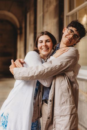 Two young people share a warm embrace, smiling joyfully, in front of a classic stone building.