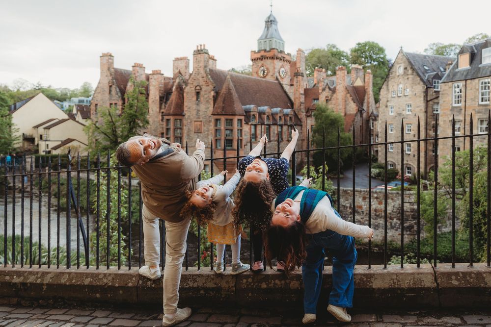 Three children playfully leaning over a railing at Dean Village in Edinburgh, Scotland, with historic stone buildings behind.