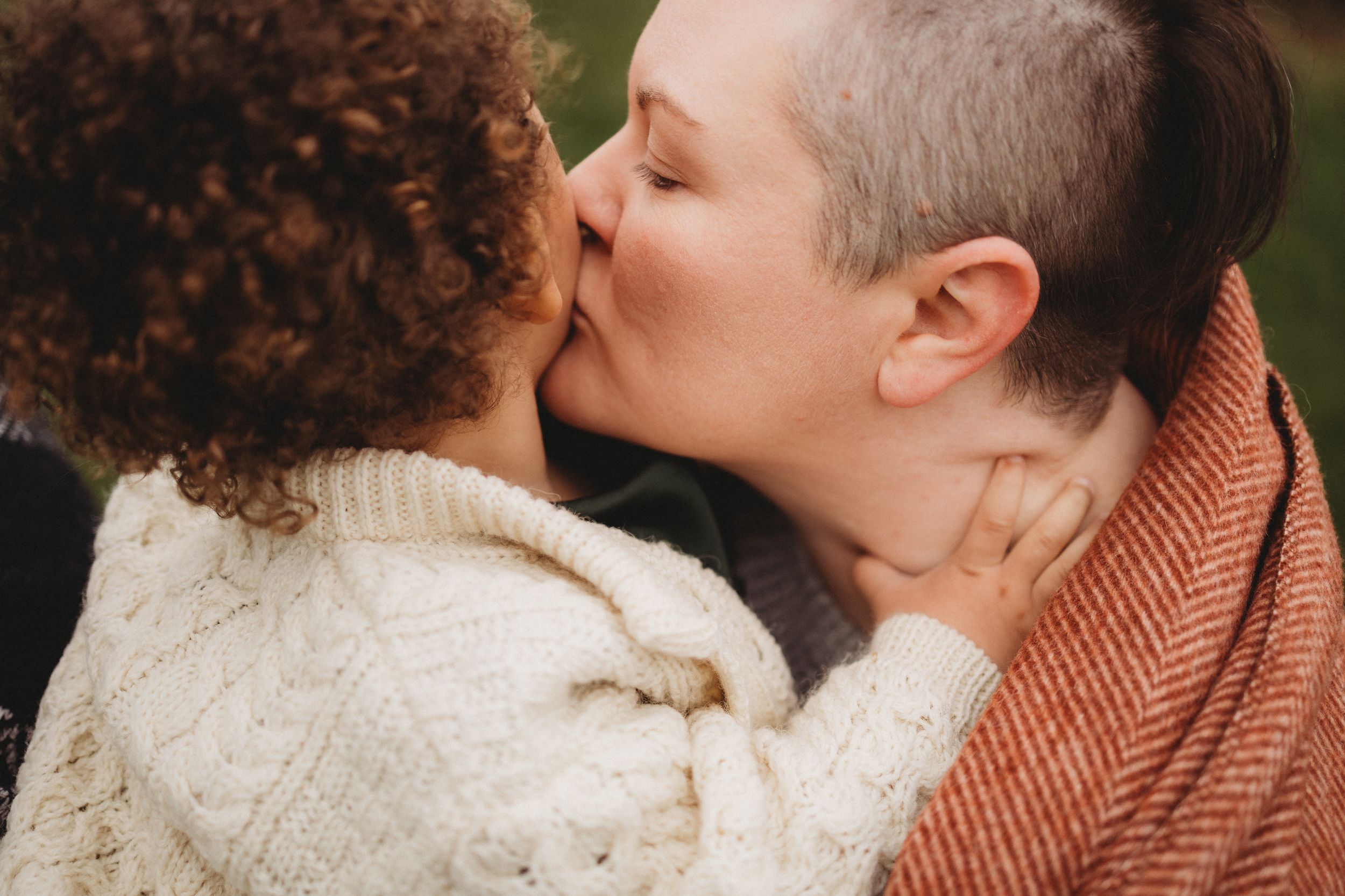 Two people sharing a tender kiss outdoors, one with curly hair in a white sweater, the other with short gray hair in an orange scarf.