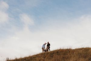 Two people stand together on a grassy hilltop silhouetted against a light blue cloudy sky.