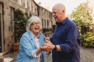 Elderly couple laughing and holding hands outdoors in a courtyard, enjoying a joyful moment together.